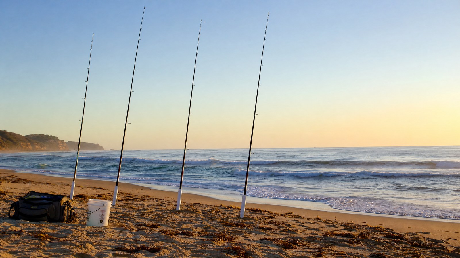 Long surf fishing rods set in sand spike holders on a beach at sunrise.