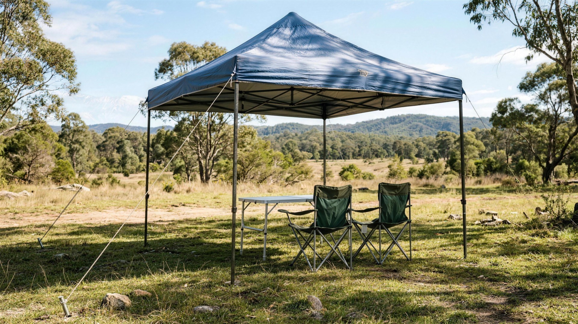 A brand-free pop-up camping gazebo shading a folding table and two camp chairs, guy-roped and pegged into open grassland