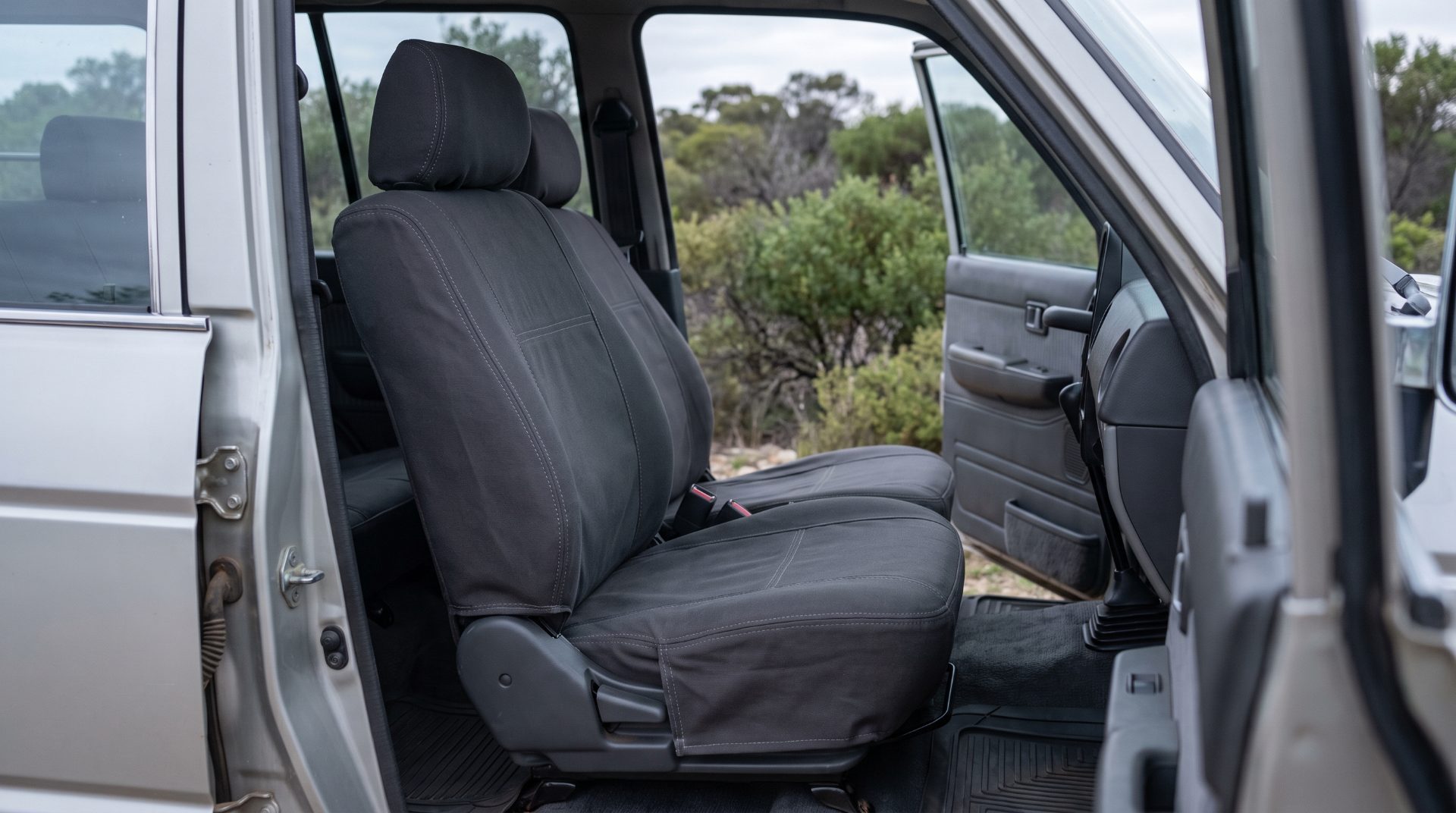 A front vehicle seat fitted with a taut dark canvas seat cover, photographed through the open door with low green scrub behind